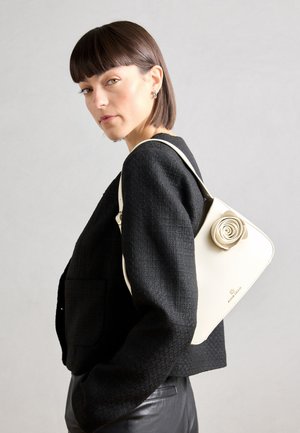 Beige handbag with rose-shaped accent, smooth texture. Worn over the shoulder, paired with a black textured jacket.