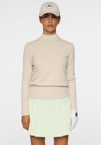Maglione beige a maglia con orlo a costine, abbinato a una gonna plissettata verde chiaro. Un berretto beige con logo e un guanto da golf bianco sulla mano sinistra.