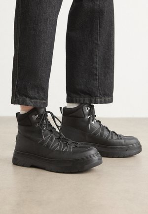 Baskets montantes noires en matériau synthétique, dotées de panneaux texturés, de semelles épaisses et de lacets avec œillets en métal. Design minimaliste.