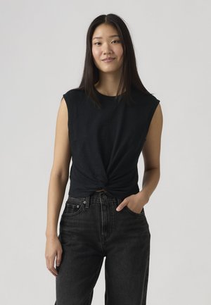 Giovane donna asiatica con capelli lunghi e neri che indossa un top nero senza maniche con nodo frontale e jeans a vita alta neri, in piedi con una mano in tasca.