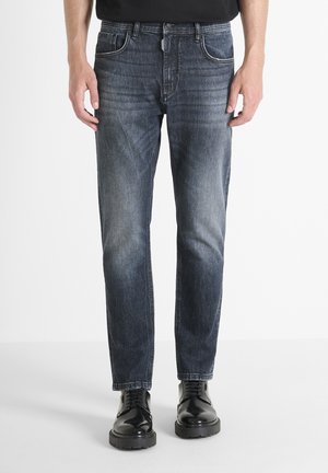 Jean en denim bleu foncé avec une coupe droite, style cinq poches, texture délavée et accents cousus aux coutures, associés à des chaussures noires.