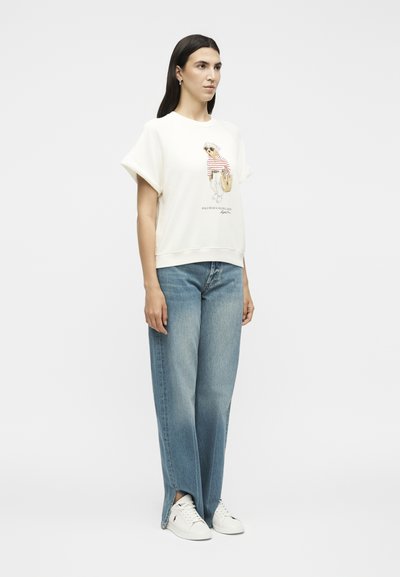 Γυναίκα φορούν λευκό κοντομάνικο φούτερ με γραφικό σχέδιο, μπλε φαρδιά τζιν με πλαϊνές σχισμές και λευκά αθλητικά παπούτσια, όρθια στο πλάι.