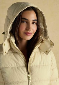 Béžová puffová bunda so lesklým zlatým zipsom a kovovými prvkami, s kapucňou a prešívanou textúrou, oblečená modelom na neutrálnom pozadí.