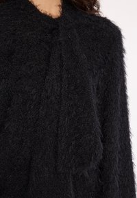 Schwarze, fuzzige Strickjacke mit einer strukturierten Oberfläche, die einen weichen, lockeren Schnitt und einen kaskadierenden Ausschnitt aufweist. Keine sichtbare Hardware oder Muster.