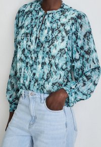 Kevyt, kuviollinen paita teal-värissä ja mustassa, jossa on pyöreä kaula-aukko, solmukuvio ja pitkät hihat. Yhdistetty korkea vyötäröinen vaaleansininen farkku.