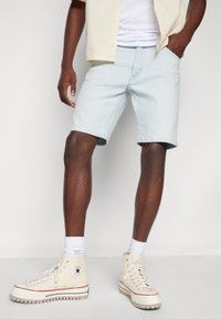 Světle modré denimové šortky s volným střihem, ohrnutým lemem a předními kapsami, doplněné béžovými vysokými teniskami a bílými ponožkami.