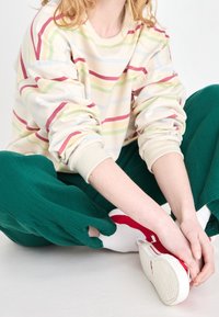 Gestreepte trui in crème, groen, roze en blauw, gecombineerd met losse groene broek. Witte sneakers met rode accenten. Casual, comfortabele pasvorm.