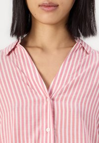 Frau mit glattem schwarzen Haar, die ein rosa-weiß gestreiftes Hemd mit offenen Kragen trägt.