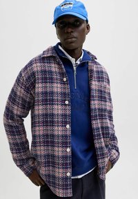 Tamsiai mėlynas, raudonas ir baltas languotas marškinėlis su mygtukais, uždėtas ant mėlyno užtrauktuko megztinio, viršuje su šviesiai mėlynu kepurėle.