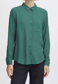 Groene blouse met lange mouwen, gemaakt van soepel materiaal, voorzien van een borstzak en verstelbare manchetten, met een gebogen zoom.