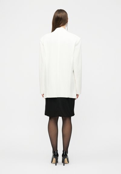 Γυναίκα στέκεται πλάτη φοράει λευκό σακάκι, μαύρη φούστα, διάφανα μαύρα καλσόν και μαύρες ψηλοτάκουνες γόβες μπροστά σε λευκό φόντο.