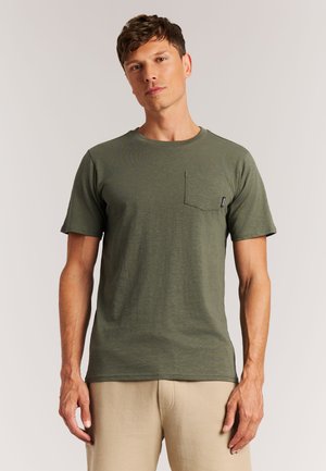 Man draagt een olijfgroen T-shirt met korte mouwen en borstzak, en beige broek, staand tegen een effen lichte achtergrond.