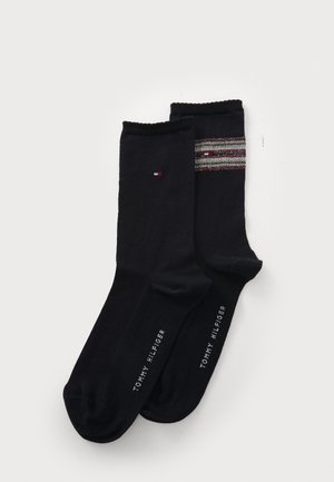 Dva páry černých ponožek leží na plocho, jeden s mnohobarevným pruhovaným vzorem. Oba mají na spodní straně vytištěné logo "Tommy Hilfiger".