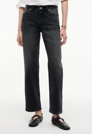 Personne portant un jean noir taille haute à jambes droites, des chaussures noires à brides, une chemise blanche et une montre-bracelet en or avec un bracelet métallique.
