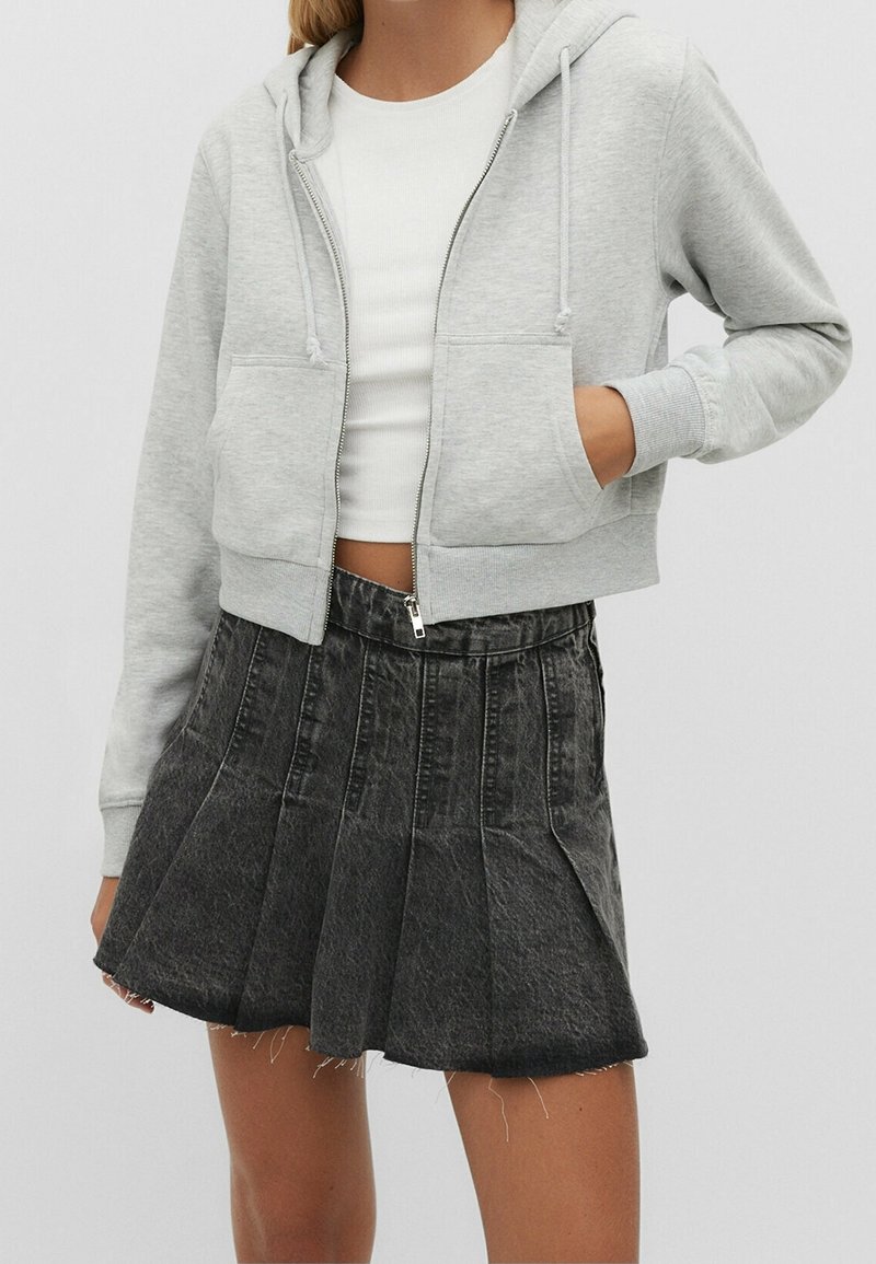 Sudadera con cremallera de color gris claro y diseño corto, con dos bolsillos frontales, combinada con una falda de mezclilla negra con pliegues y detalles de dobladillo sin acabar.