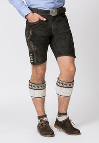 Shorts in suede scuro con dettagli ricamati, tasche laterali e bottoni decorativi. Abbinati a scaldamuscoli bianchi in maglia e scarpe marroni.