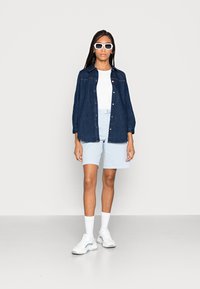 Camicia di jeans blu scuro, t-shirt bianca aderente, pantaloni corti azzurri, scarpe da ginnastica bianche con accenti blu e occhiali da sole bianchi. Sfondo semplice.