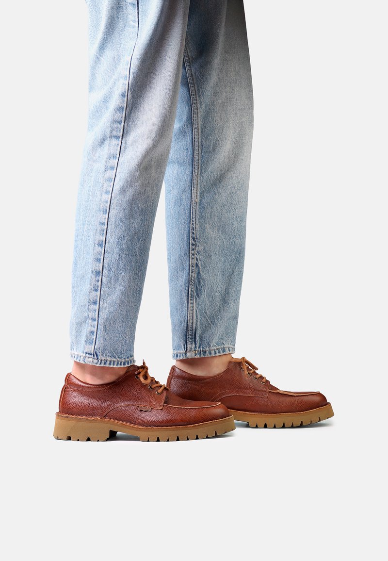 Personne portant un jean bleu clair et des chaussures en cuir marron à lacets avec des semelles épaisses beige clair, debout sur fond blanc.