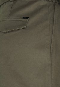 Olivově zelená látka s hladkou texturou, zdobená našitým kapsou a jemným kovovým detailem na boku.