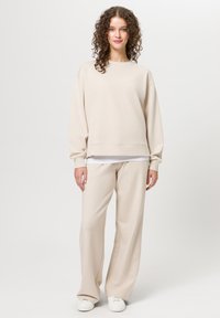 Sweatshirt beige chiaro con scollo rotondo e spalle scese, abbinato a pantaloni a gamba larga. Entrambi realizzati in tessuto morbido, con dettagli minimi.