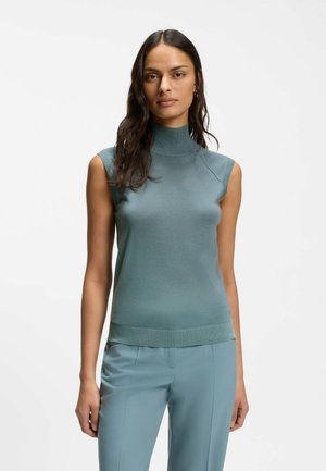 Donna con capelli lunghi e scuri che indossa una maglia smanicata a collo alto color verde acqua e pantaloni abbinati sullo sfondo bianco semplice.