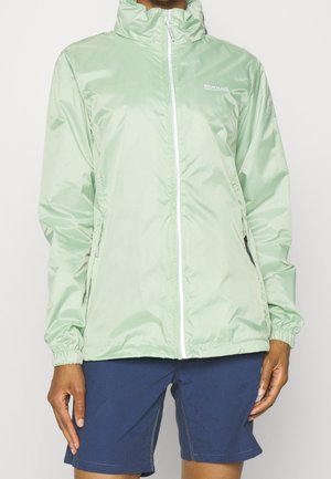 Person trägt hellgrüne Windjacke mit Reißverschluss und dunkelblaue Shorts, steht vor einfachem Hintergrund.