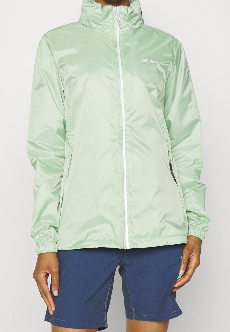 Personne portant une veste coupe-vent zippée vert clair et un short bleu foncé, debout devant un fond uni.