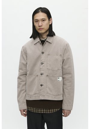 Lys beige bomuldsjakke med tre frontlommer, knaplukning og et mærke på siden. Bæres over en brun sweater og en skovmandsskjorte.