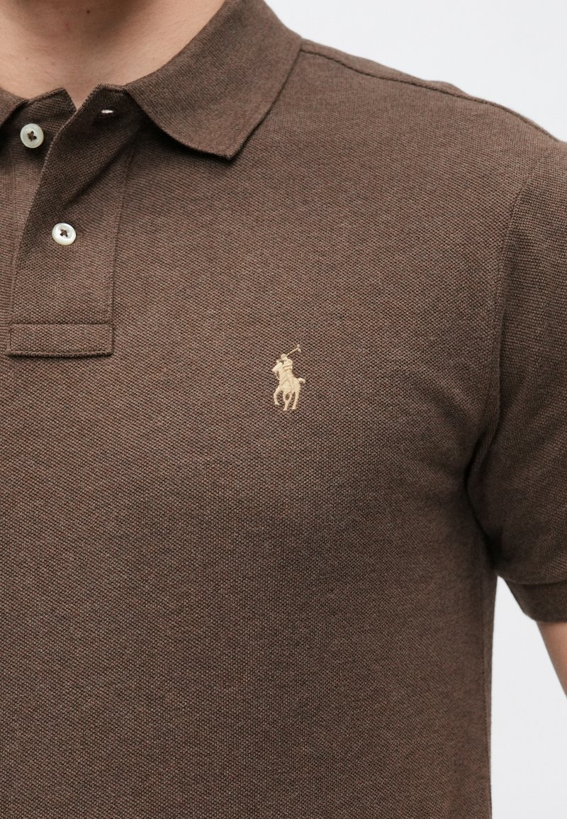Braunes Poloshirt aus strukturiertem Material. Verfügt über einen klassischen Kragen, zwei Knöpfe und ein goldfarbenes, gesticktes Logo auf der linken Brust.