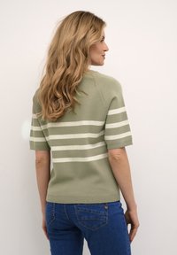 Kortærmet olivengrøn sweater med hvide vandrette striber; blød tekstur og ribbet kant. Båret med blå jeans, som viser et afslappet look.
