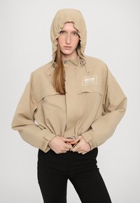 Bežinė striukė su gobtuvu, priekyje su užtrauktuku ir virvutėmis; turi Moschino Jeans logotipą ir laisvus rankovės.
