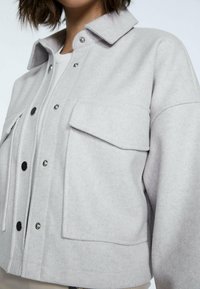 Szara kurtka z wełny o wyglądzie wełny, z ostro zakończonym kołnierzem, zatrzaskami, dwoma kieszeniami na piersi i luźnym krojem. Gładka tekstura i minimalistyczne detale projektu.
