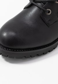 Black leather boot featuring a textured surface, rounded toe, and lugged rubber sole. Includes eyelets for laces and a stitched trim.