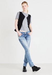 Un gilet sans manches noir sur un t-shirt gris. Jean droit délavé bleu clair. Bottes noires à lacets. Fond blanc.