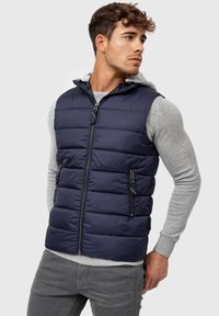 Marineblå puffervest med lynlås foran, to sidelommer og hætte, lagdelt over en lys grå langærmet skjorte. Slankt, quiltet design.