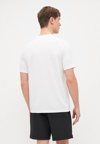 Kurze weiße Sportshirt mit glatter Textur, entworfen für Komfort. Kombiniert mit schwarzen strukturierten Shorts, Rückansicht.