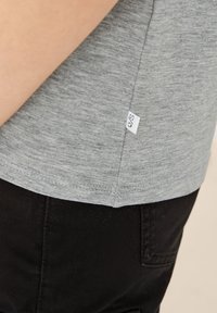 Szary t-shirt z krótkim rękawem o gładkiej teksturze, z małą białą metką z boku. Połączony z czarnymi spodniami.