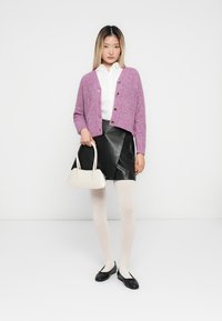 Rosa strikket cardigan med knapper, hvit skjorte, svart skinnskjørt, gjennomsiktige strømpebukser og svarte leiligheter. Holder en liten lysbeige håndveske.