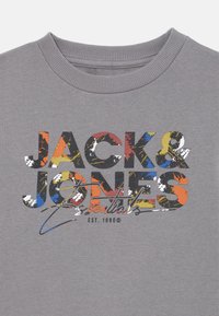 Pilkas sportinis megztinis su raištiniu apykaklės dizainu. Pasižymi spalvinga grafine spausdinta „JACK & JONES Essentials“ įvairiais šriftų stiliais.