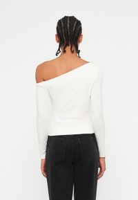 Dolga majica z odkrito rameni, bela, izdelana iz mehkega materiala. Prilagojena oblika z ukrivljenim izrezom, v kombinaciji s črnimi hlačami z visokim pasom.