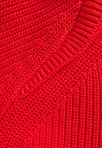 Sweter robiony na drutach w jasnej czerwieni, z różnymi fakturami, posiadający żebrowany wzór oraz gładki, luźny splot.