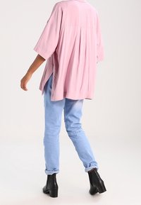 Rosa Bluse mit kurzen Ärmeln und Rückenschlitz-Detail, kombiniert mit hellblauen Jeans mit hochgekrempelten Ärmeln und schwarzen Knöchelstiefeln. Glatte Stoffoberfläche.