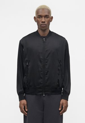 Jeune homme aux cheveux blonds courts portant une veste bomber noire zippée et un pantalon sombre, debout devant un fond blanc uni.