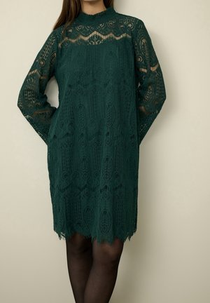 Vrouw draagt een donker groene kanten jurk met lange mouwen, knielengte, hoge halslijn en doorschijnende patroon secties op de mouwen en borst.