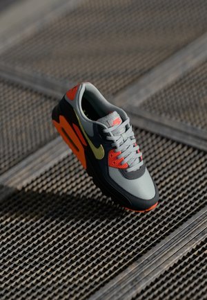 Nike skriešanas apavi pelēkā krāsā ar oranžiem un melniem akcentiem. Iekļauj acs un ādas augšdaļu, spilventotu apkakli un teksturētu zoli.