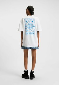 Witte oversized t-shirt met een blauwe grafische print van hemelse elementen en handen op de achterkant, gecombineerd met een denim rok en zwarte laarzen.