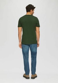 Man wearing dark green t-shirt, blue jeans, and brown shoes, standing facing away against a plain light background.