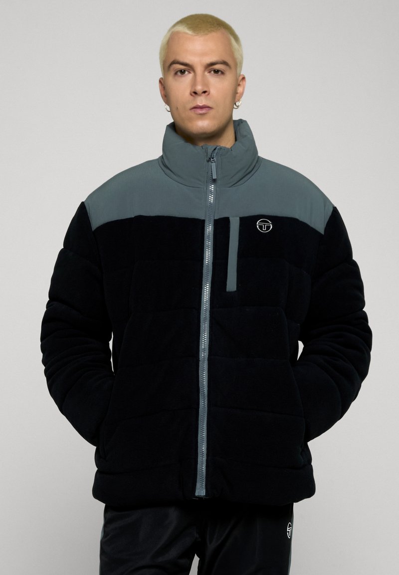 Pufferjakke med svart og grå fargekombinasjon, quiltet design, høy krage og frontglidelås med liten logo på brystet.