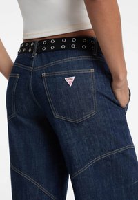 Donkerblauwe denim spijkerbroek met wijde pijpen, met topstiksel, een driehoekige rood-witte logo patch op de achterzak en een zwarte metalen gesp riem.