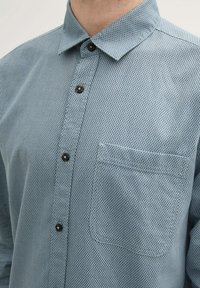 Camisa de botones en azul claro con un patrón de cuadros texturizado. Cuenta con un cuello, botones negros y un bolsillo en el pecho del lado izquierdo.